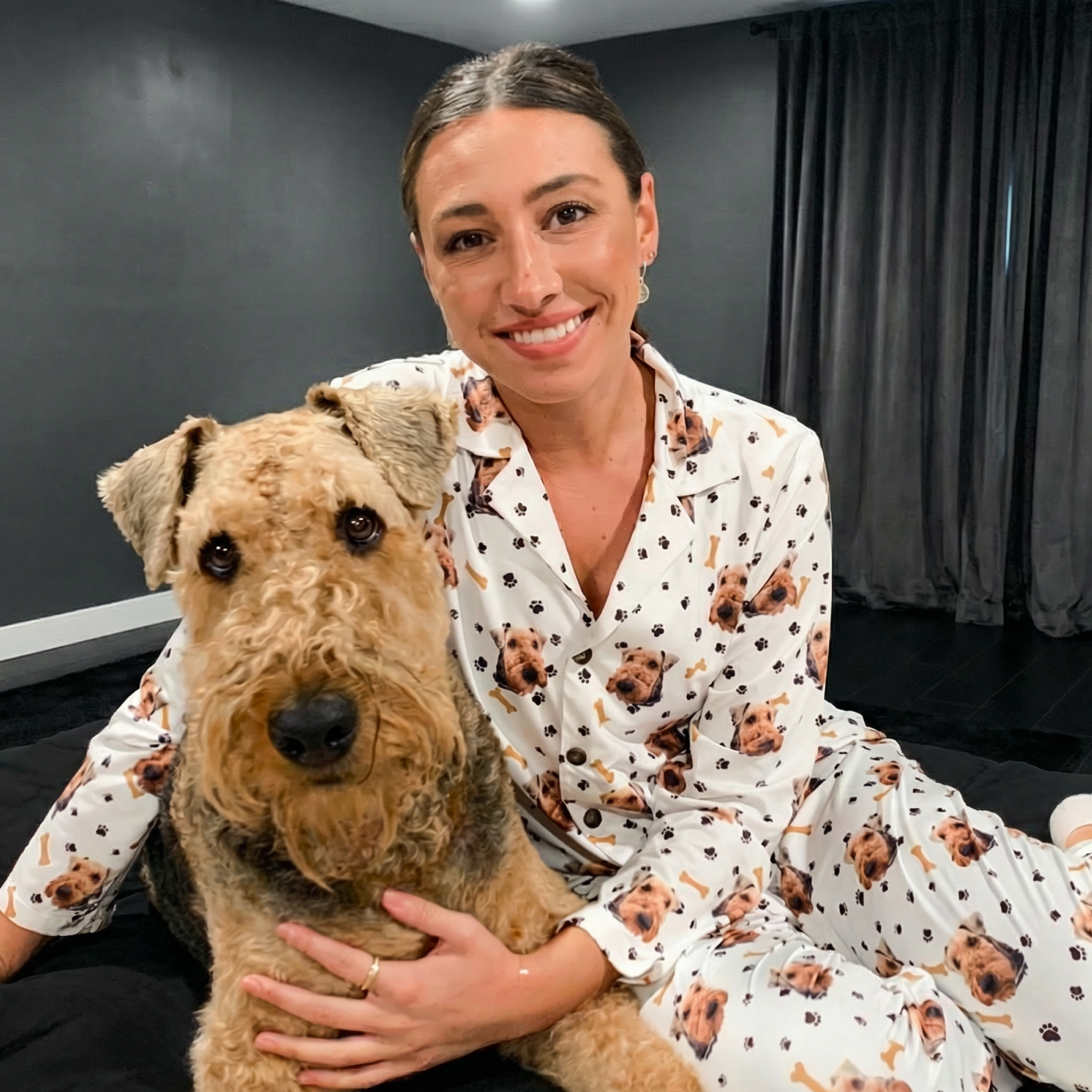 Smiling woman on bed with Airedale Terrier wearing white pajamas printed with the dog’s face and paws