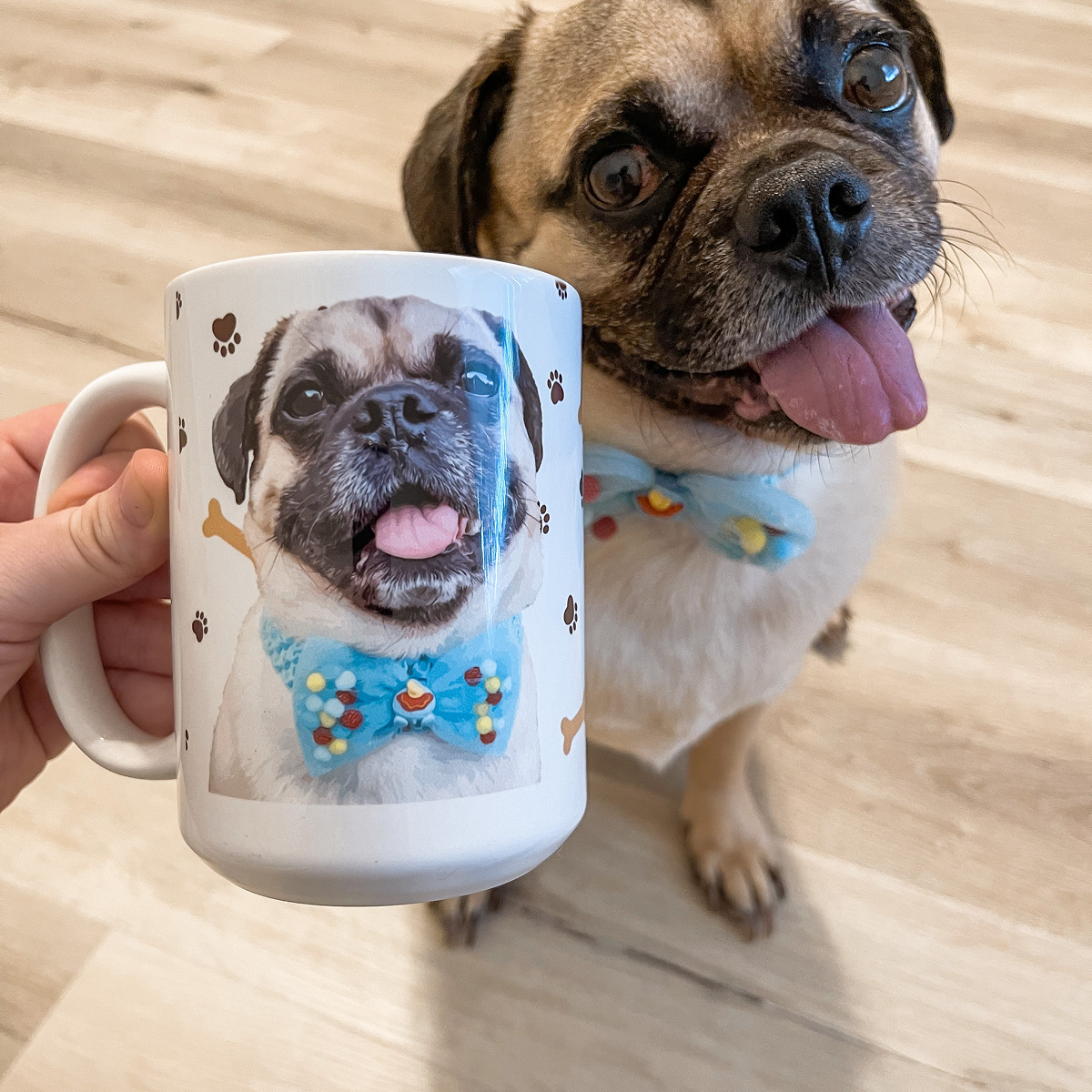 A person's hand is holding a white mug that features a portrait of a smiling Pug wearing a light blue bow tie. 