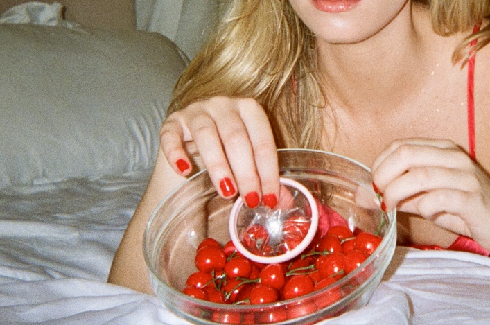 Person with red nails holding a bowl of cherries and a pink condom.