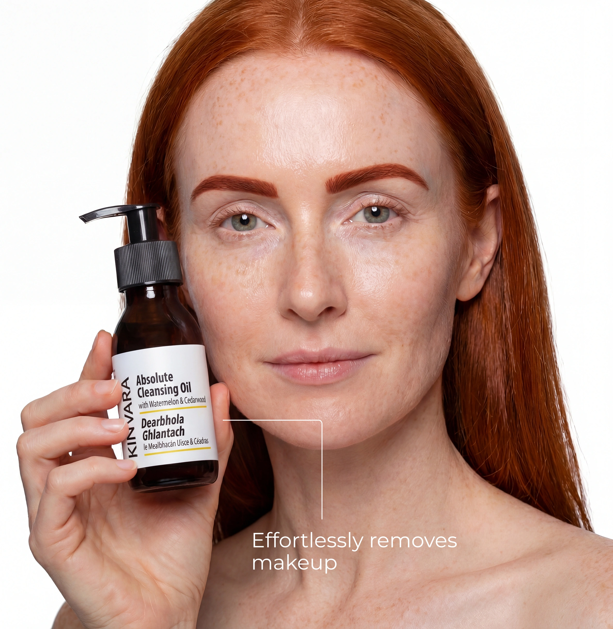 A woman with red hair and freckles holds a bottle of Kinvara Absolute Cleansing Oil.