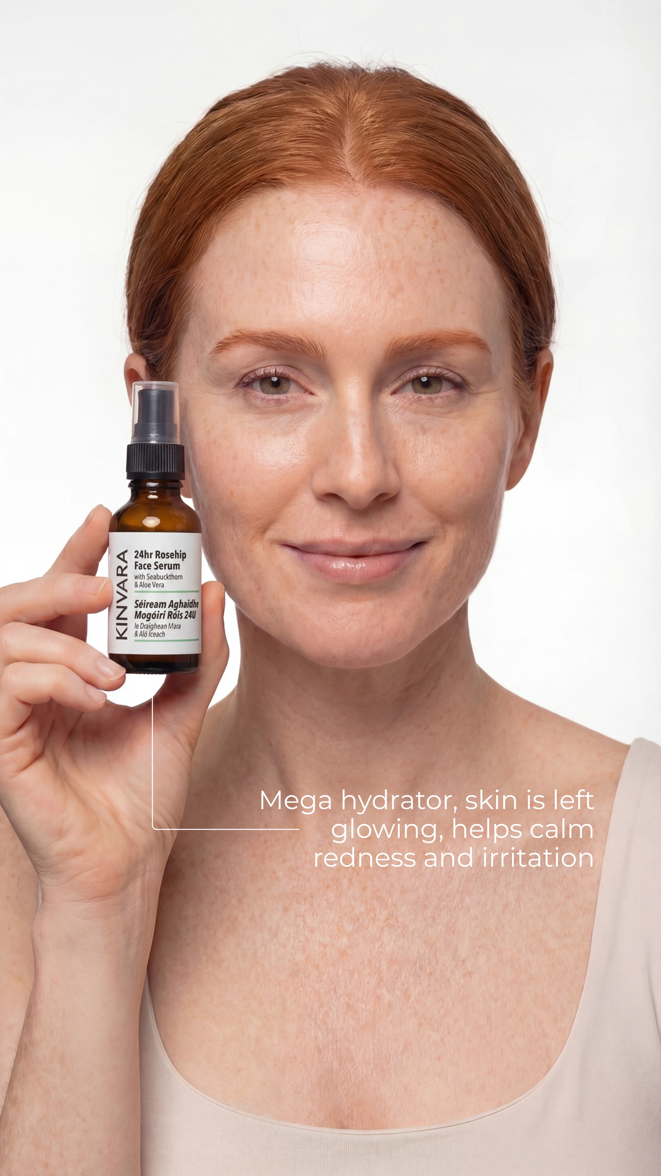 A woman with red hair and freckles smiling while holding a bottle of Kinvara 24hr Rosehip Face Serum.