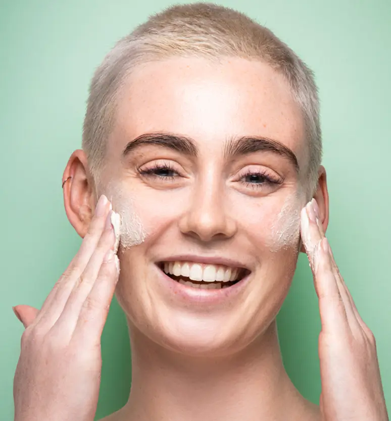 Person smiling while applying cream to their face against a green background.