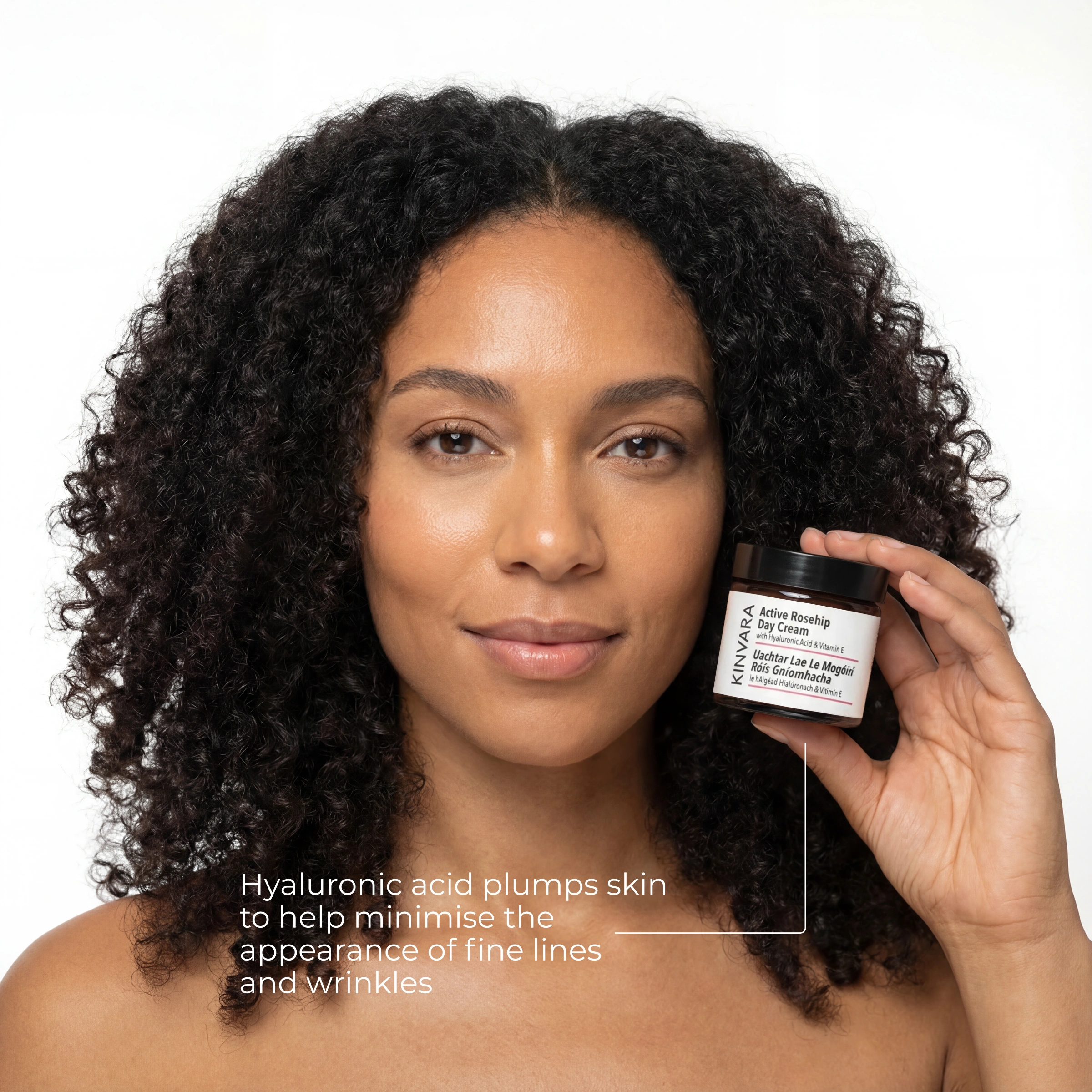A woman with dark curly hair holds up a jar of Kinvara Active Rosehip Day Cream.