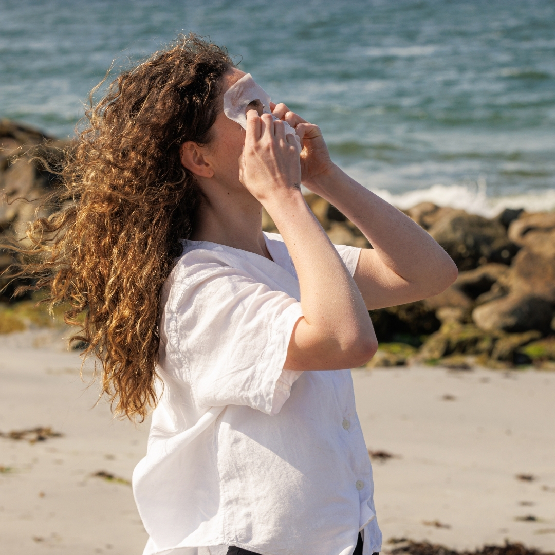 Person on beach applying a face mask, ocean in background.