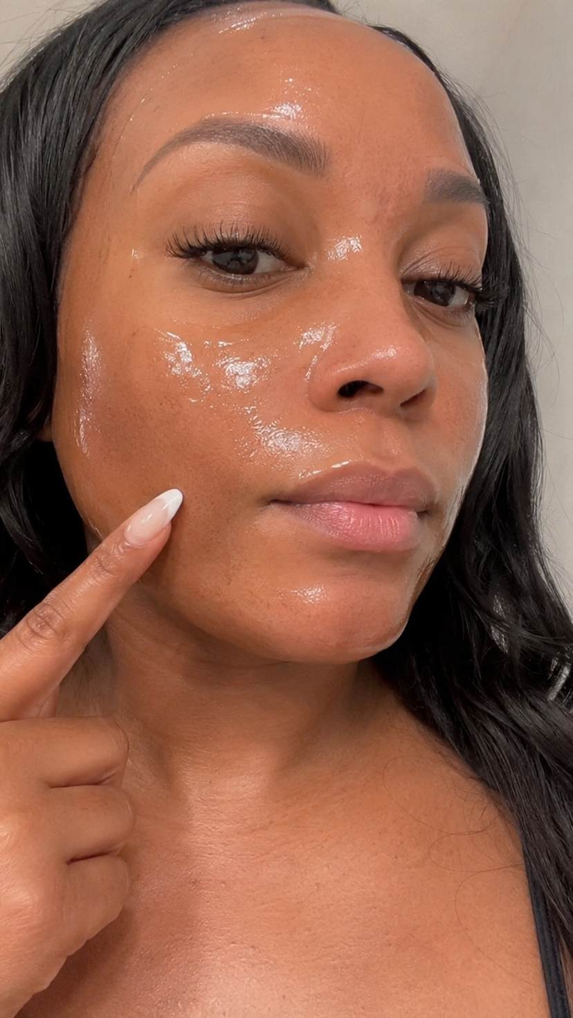A close-up of a woman with a glossy, clear product on her face pointing to her cheek.
