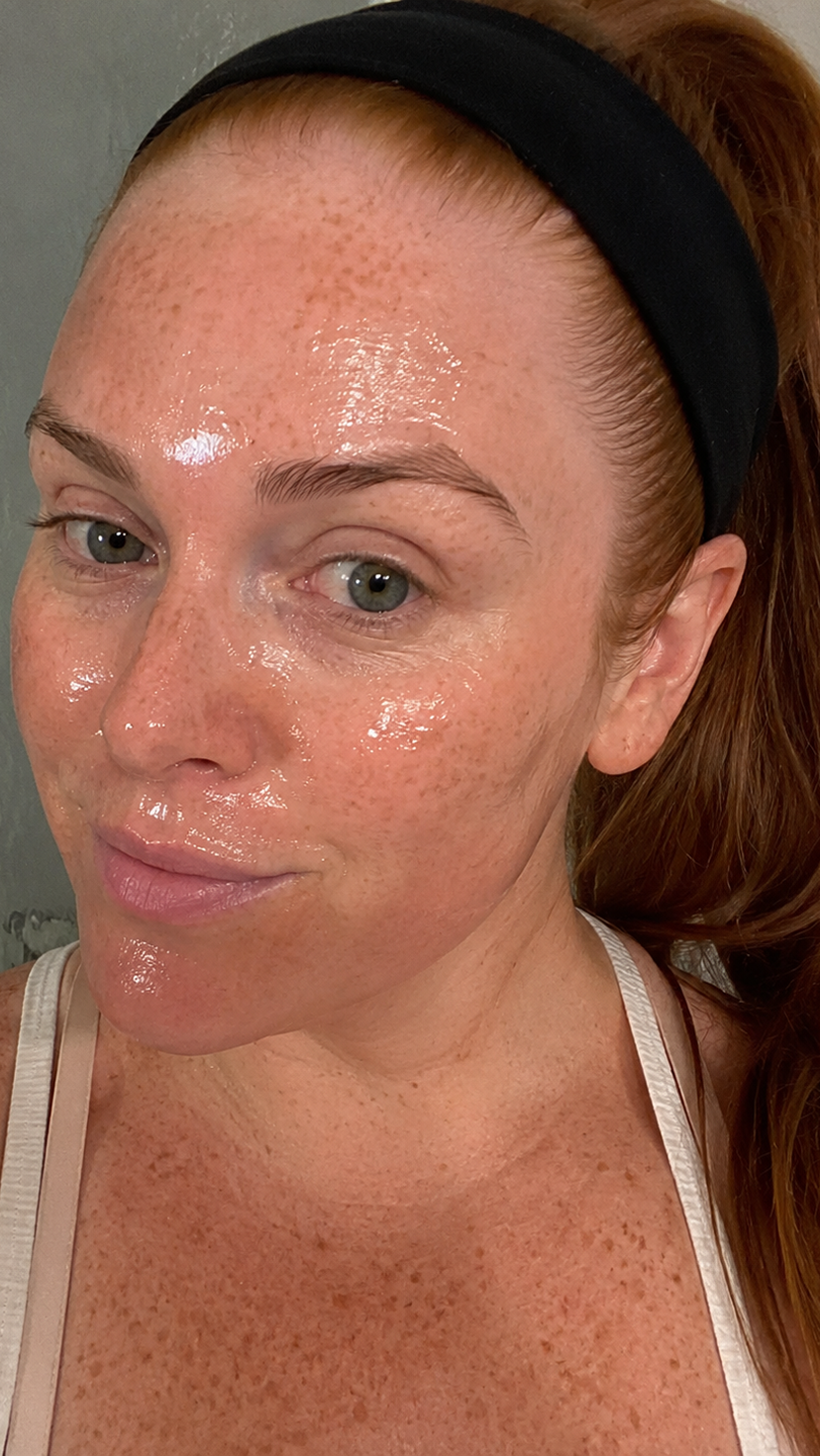 A close-up of a woman with red hair and freckles, her face covered in a shiny serum.
