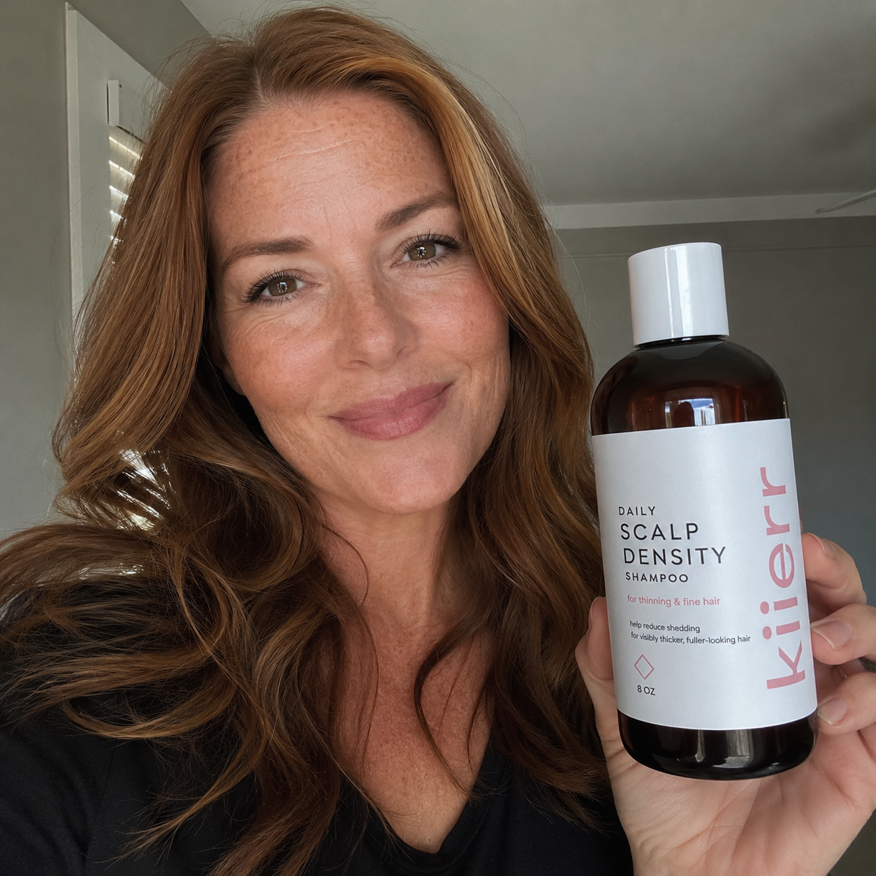 A woman with long, reddish-brown hair holds up a bottle of Kierr Daily Scalp Density Shampoo.