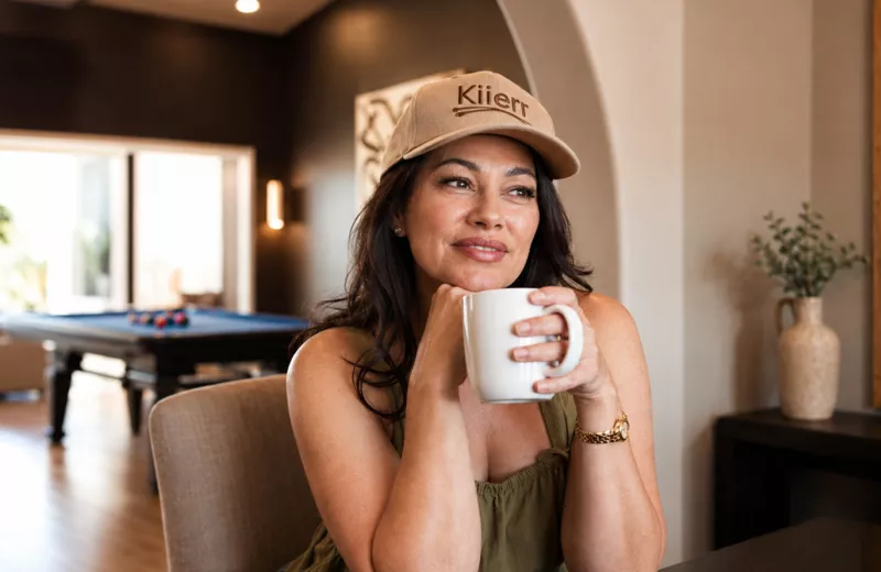 A woman in a 'Kiierr' baseball cap holds a white mug while looking off to the side.