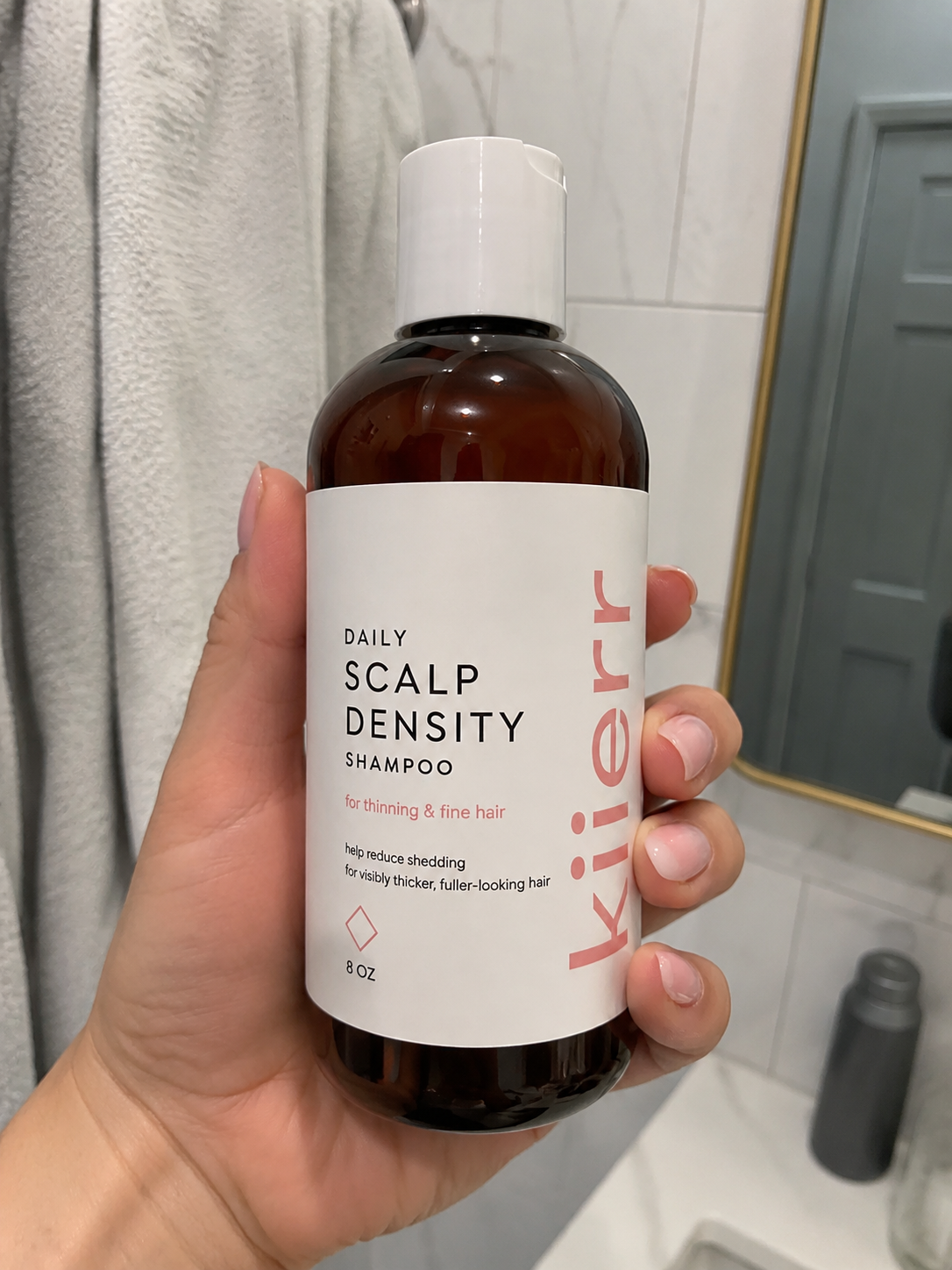 A hand holds a bottle of Kierr Daily Scalp Density Shampoo in a bathroom setting.