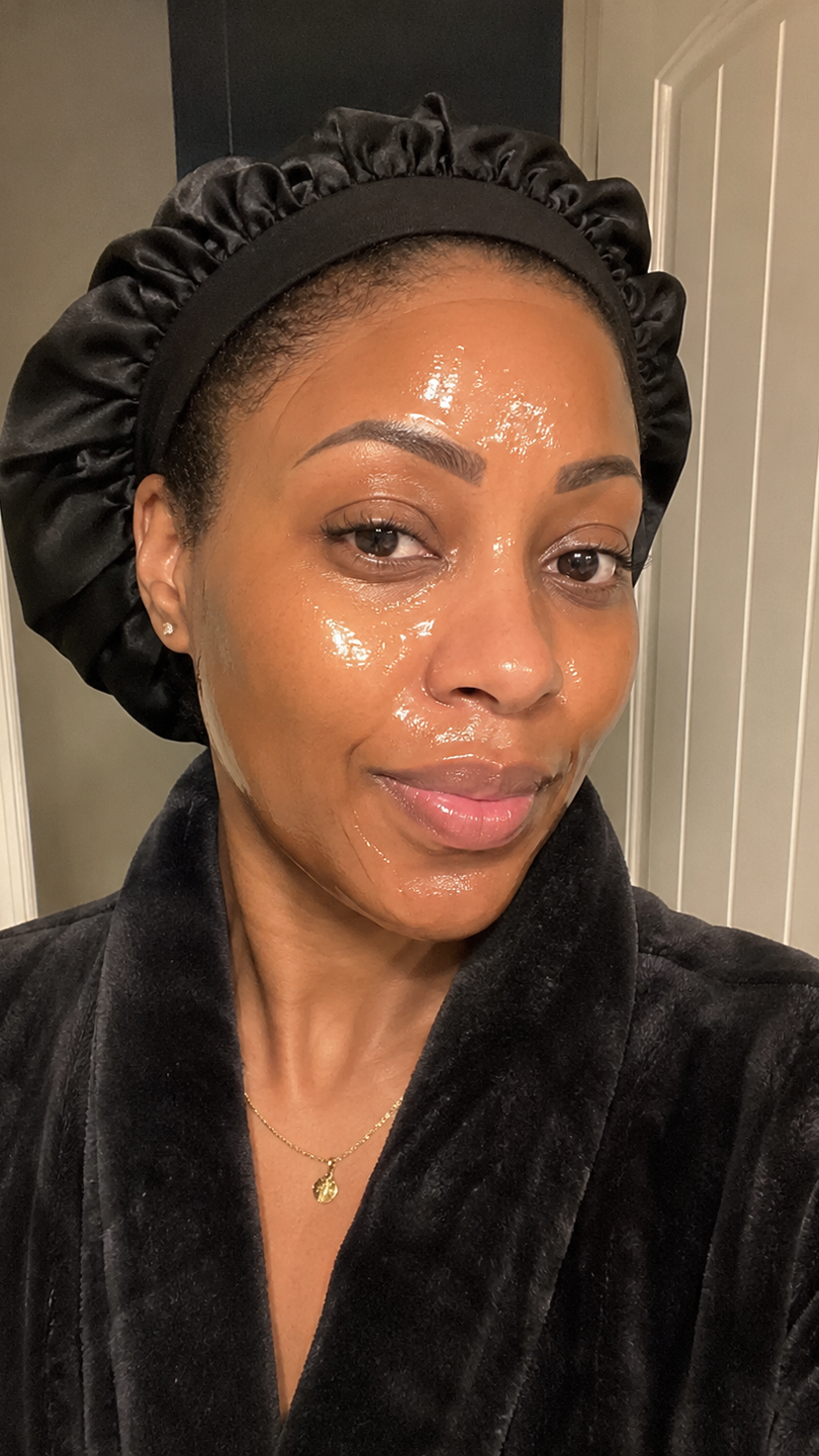 A woman in a black satin bonnet and robe with a glossy skincare product on her face.