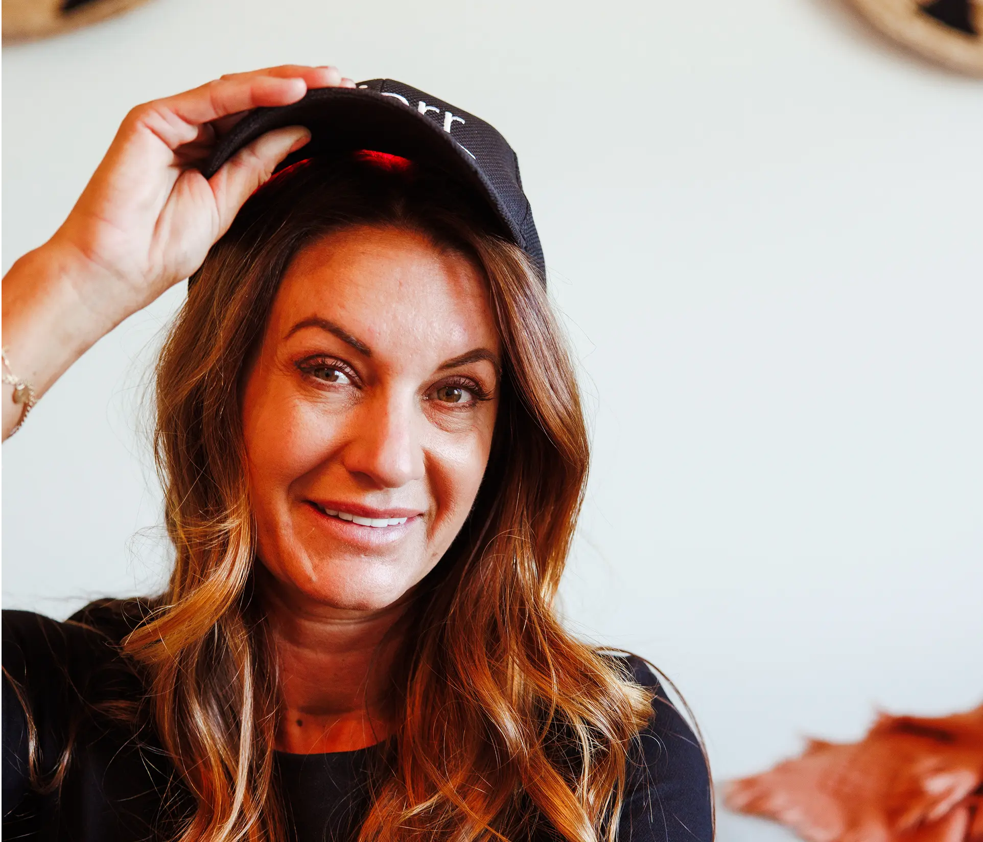 A smiling woman with long brown hair holds a black baseball cap on her head.