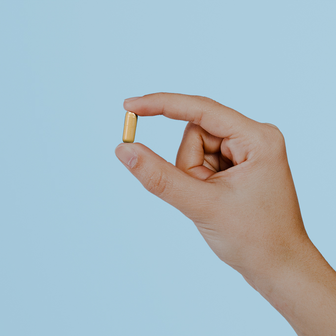 Hand holding a small yellow capsule against a light blue background.