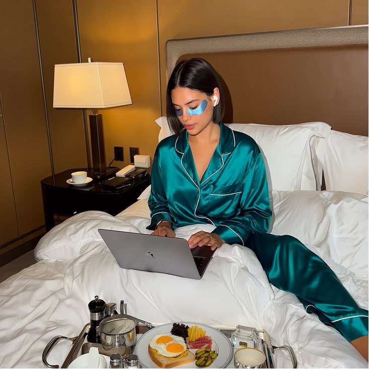 Person in teal pajamas on bed with laptop and breakfast tray.