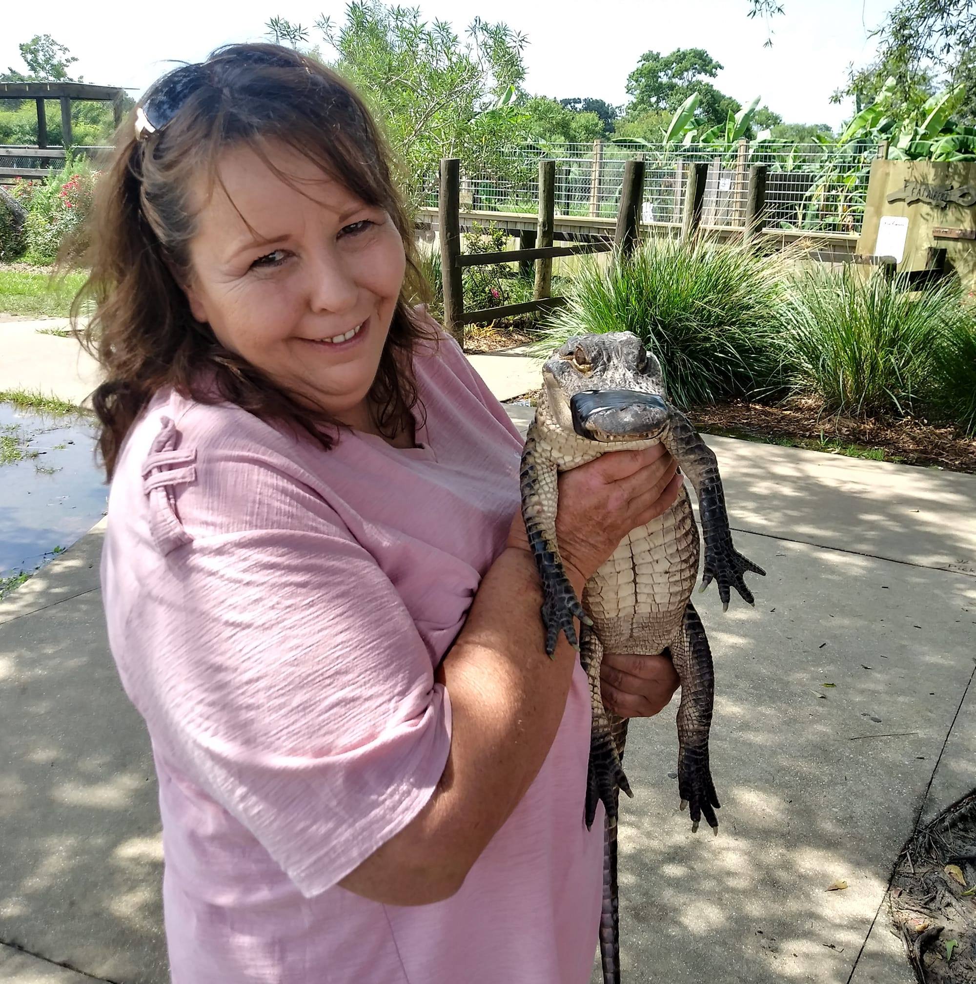 A smiling woman in a pink shirt holds a small alligator with its mouth taped shut.