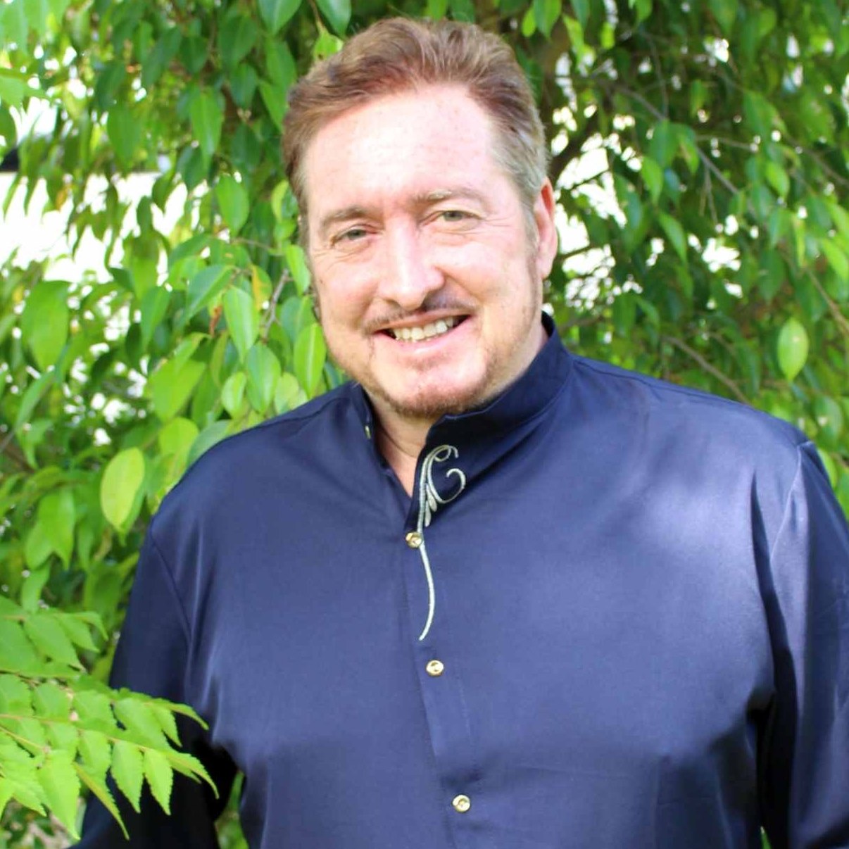 A smiling man with reddish-brown hair and a goatee, wearing a blue high-collared shirt, stands outdoors.