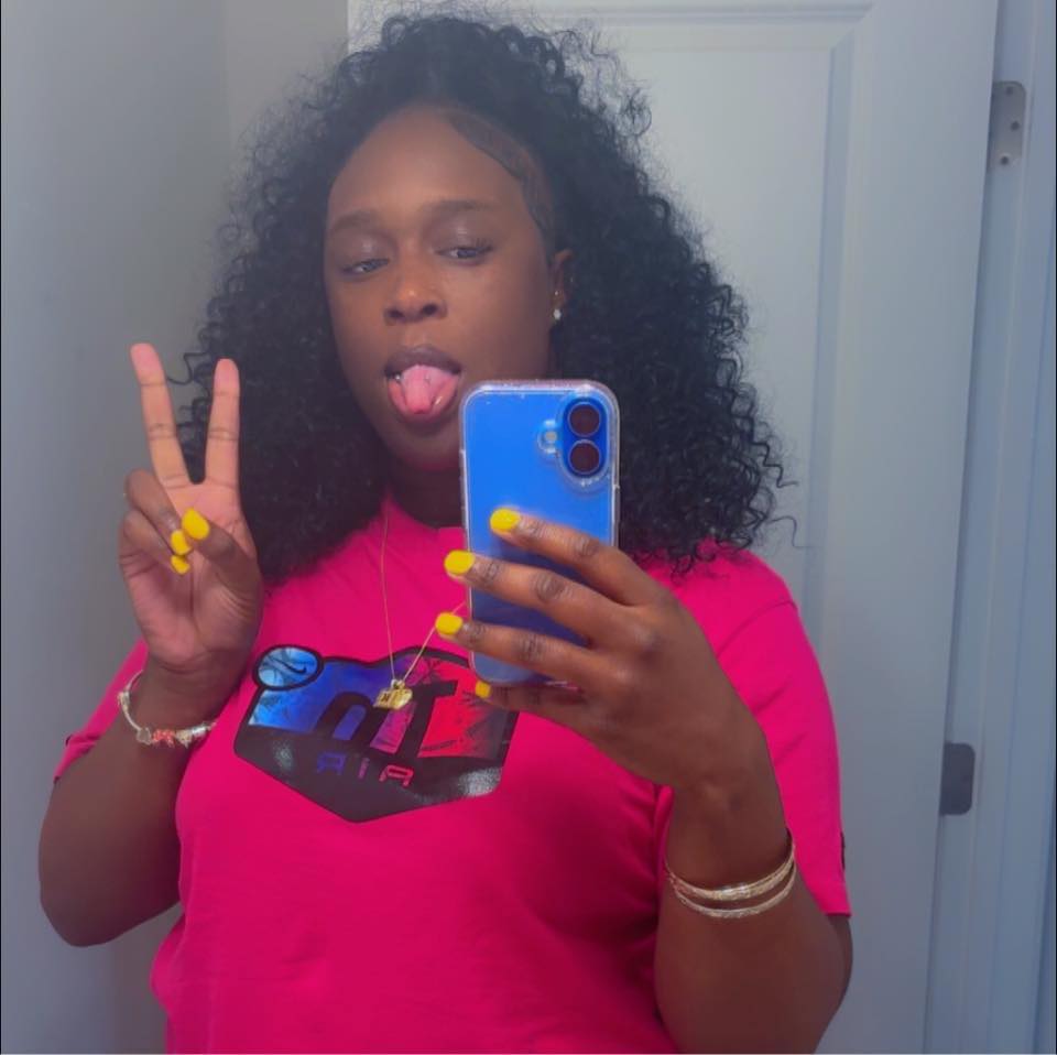 A woman in a pink shirt takes a mirror selfie, making a peace sign and sticking her tongue out.