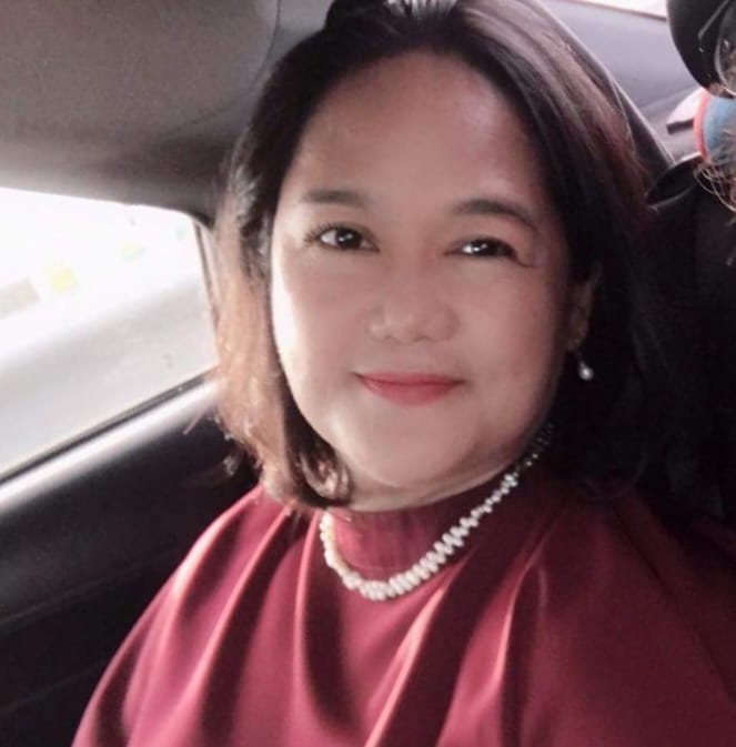 A woman with dark, shoulder-length hair and a pearl necklace smiles while sitting inside a car.