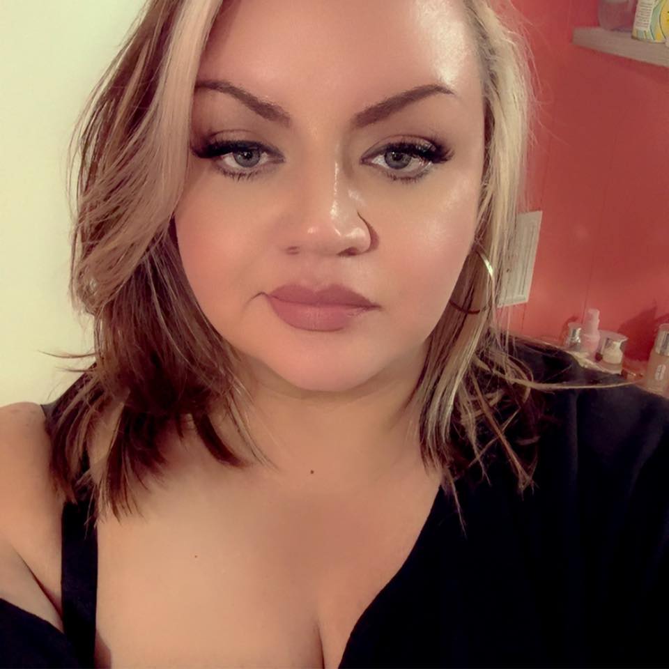 A close-up of a woman with blonde hair and makeup, wearing hoop earrings and a black top.