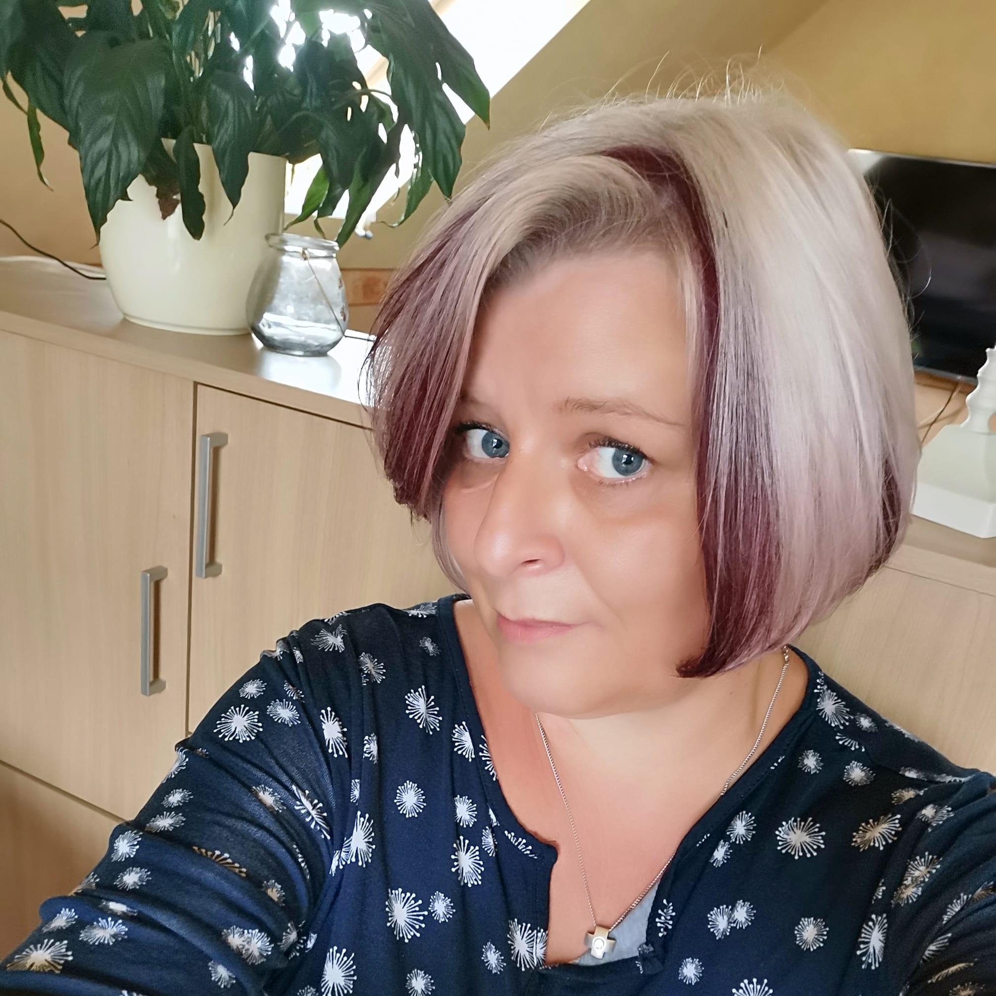 A woman with short blonde and reddish-purple hair in a patterned shirt looks at the camera.