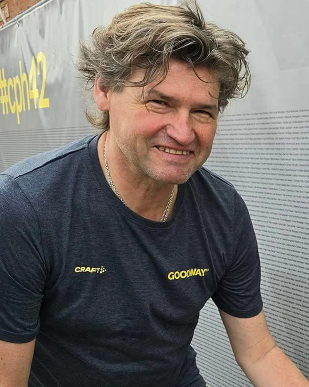 Man smiling, wearing a blue shirt with 'Craft' and 'Goodway' logos, standing outdoors.