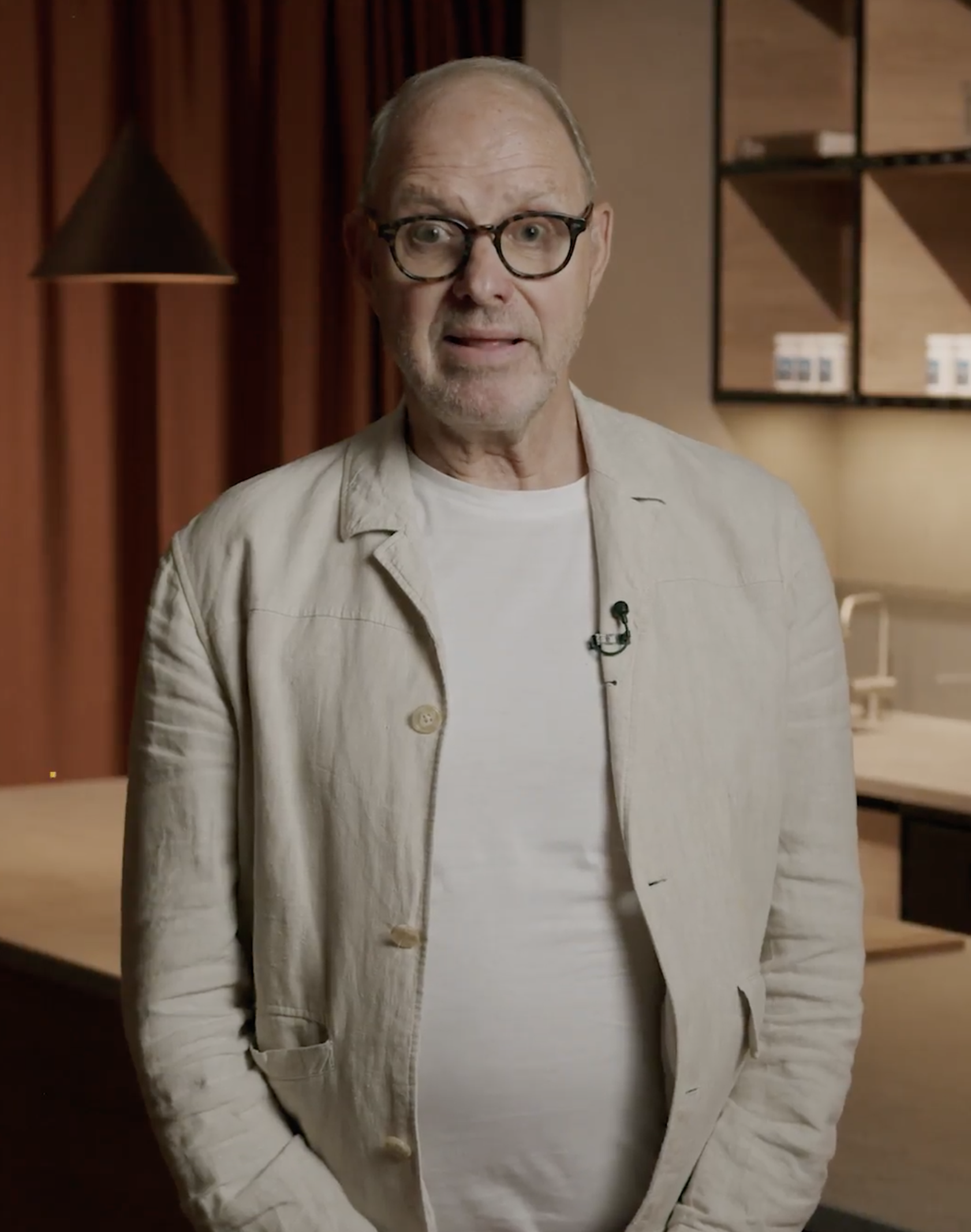 Man in glasses and beige jacket standing in a warmly lit room.