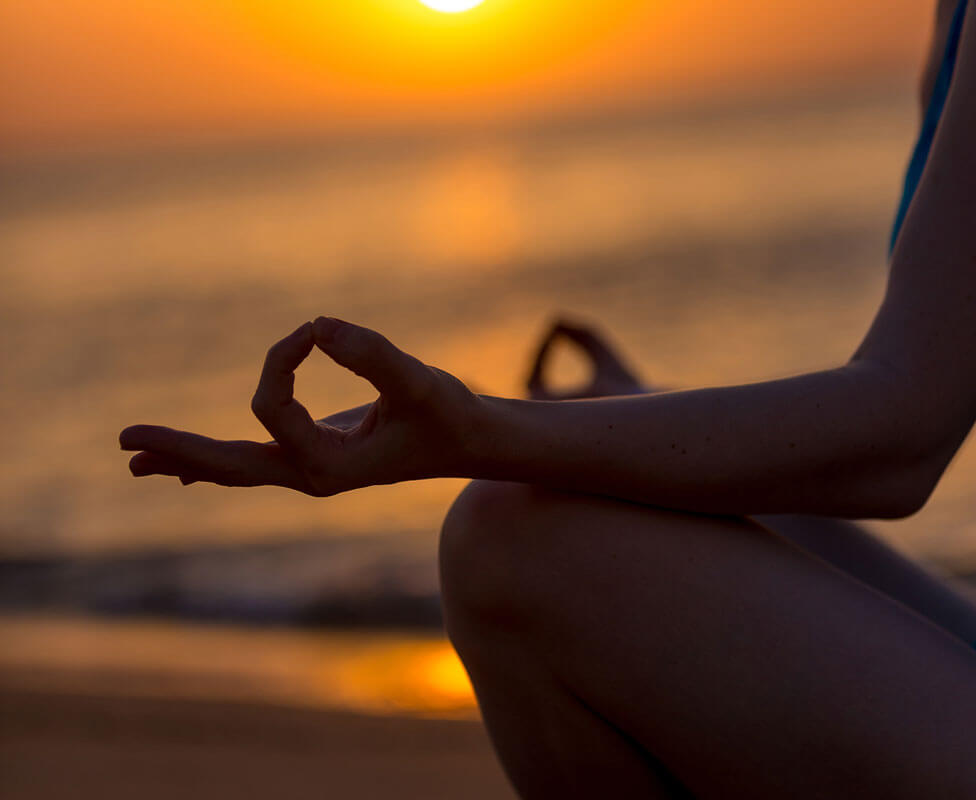 Person meditating on a beach at sunset, with hands in a mudra position.