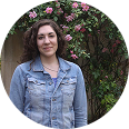 Person posing in front of a flowering bush, wearing a denim jacket.