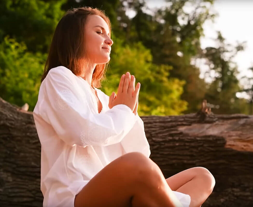 Woman meditating outdoors with hands in prayer position, surrounded by trees.