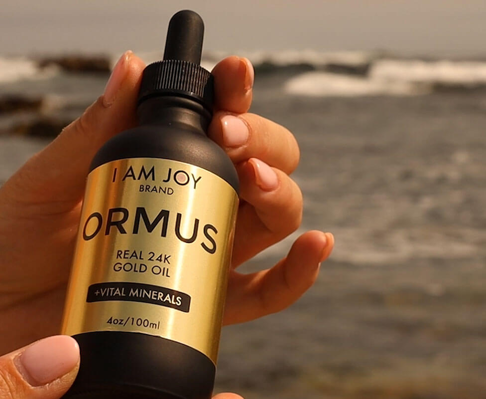 Hand holding a bottle of ORMUS gold oil by the ocean.