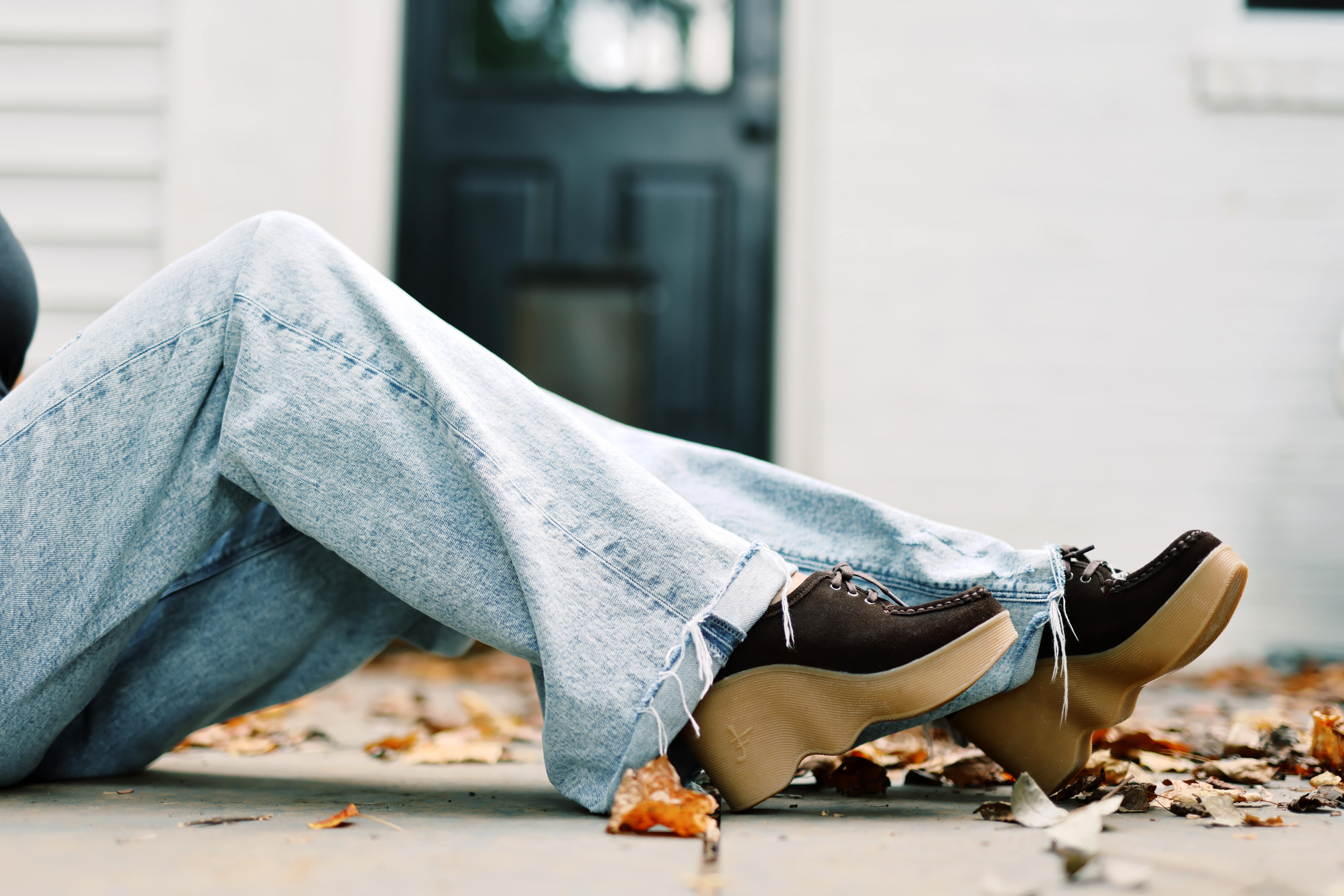 Person wearing platform shoes and frayed jeans, sitting near a black door with fallen leaves.