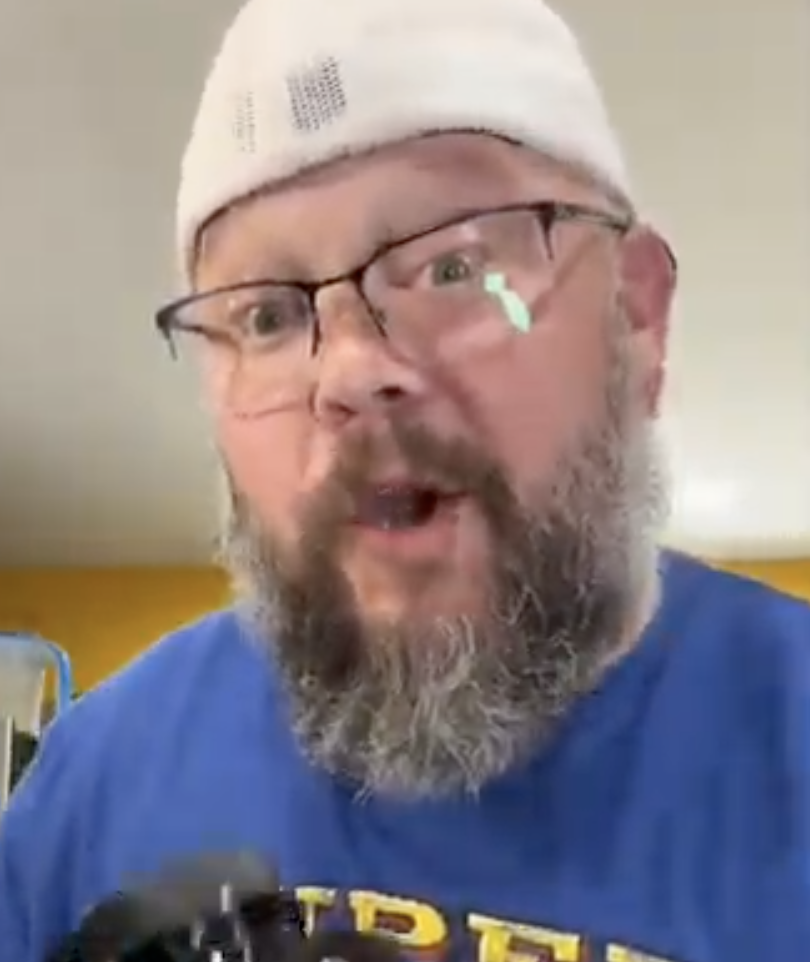 Man with glasses and a beard wearing a white cap and blue shirt.