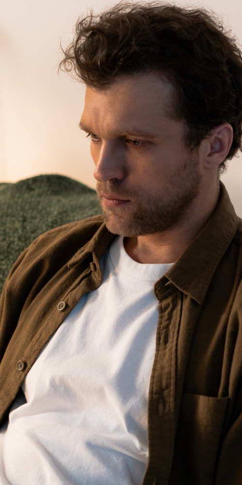 A man with a beard and curly hair, wearing a brown shirt, looking thoughtful.