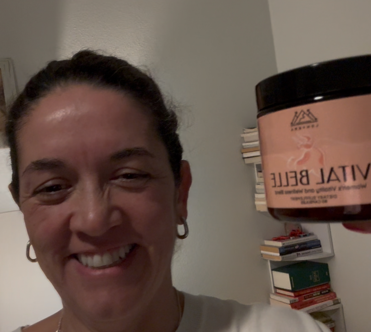 Person smiling and holding a jar labeled 'Vital Belle' in a room with books.