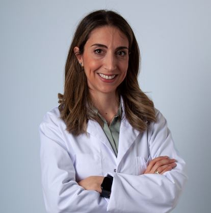 A smiling woman with long brown hair wearing a white lab coat with her arms crossed.