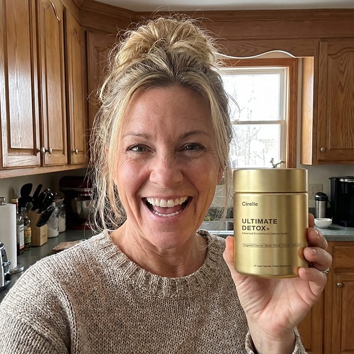 A smiling woman in a kitchen holds up a gold-colored bottle of Cirelle Ultimate Detox+ supplements.