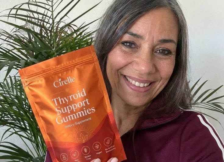 A smiling woman holds up an orange pouch of Cirelle Thyroid Support Gummies in front of a plant.