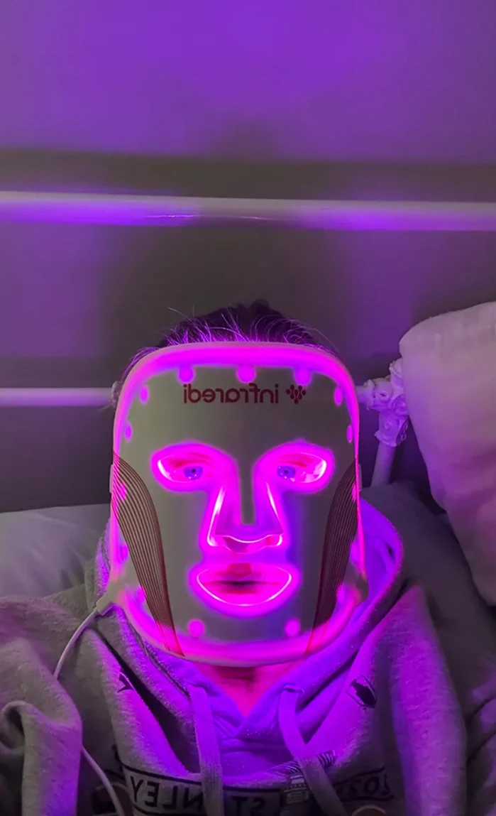 Person wearing an illuminated LED face mask, glowing purple, lying down.