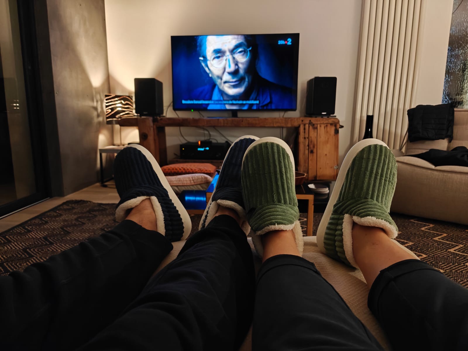 Two people with slippers relaxing in front of a TV showing a man's face.