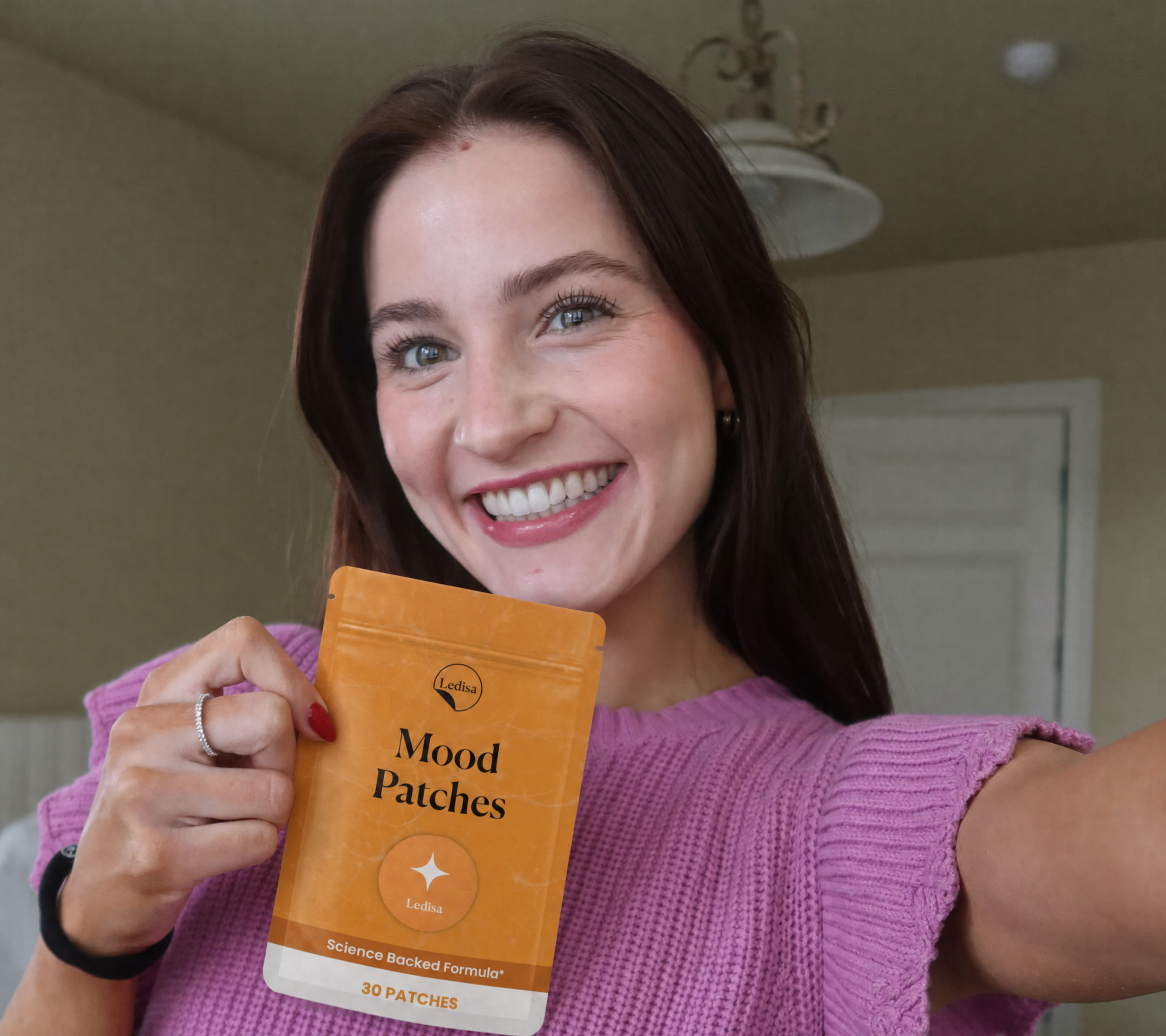 A smiling woman in a purple sweater holds up a package of Ledisa Mood Patches.
