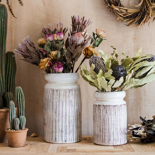 Two rustic vases with dried flowers on a wooden table, surrounded by cacti.