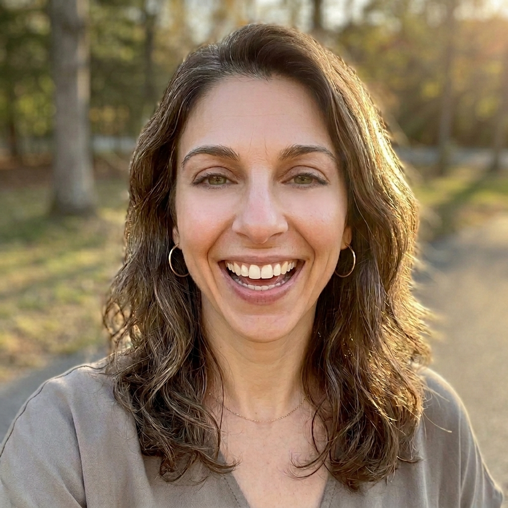 A close-up headshot of a smiling woman with wavy brown hair, standing outdoors.