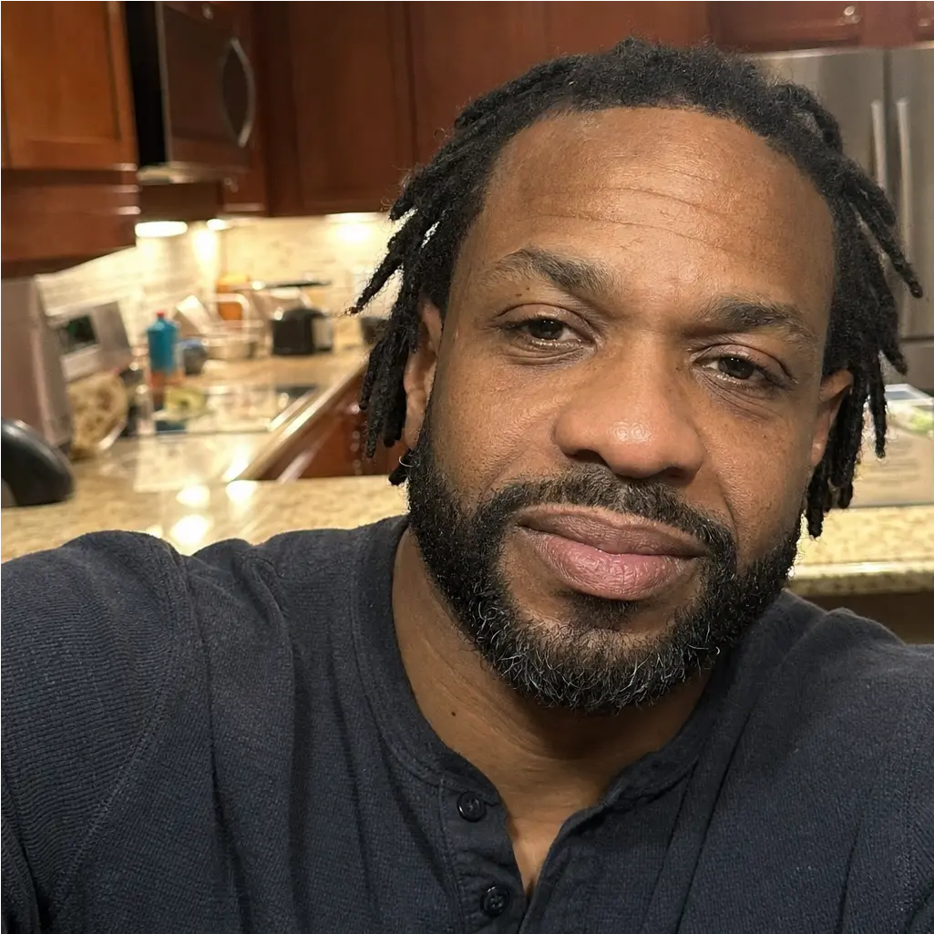 A man with dreadlocks and a beard takes a selfie in a kitchen.
