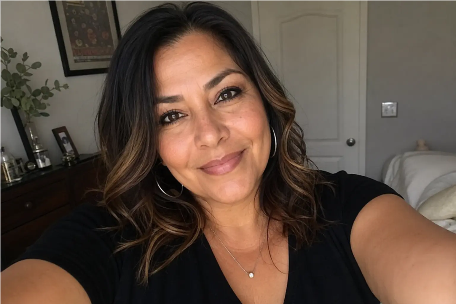 A smiling woman with wavy brown hair, wearing a black shirt and hoop earrings, takes a selfie indoors.