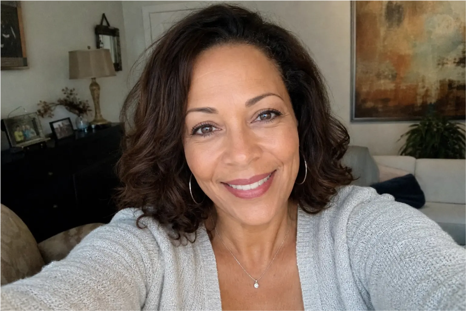 A woman with dark curly hair and hoop earrings smiles warmly at the camera from inside a home.