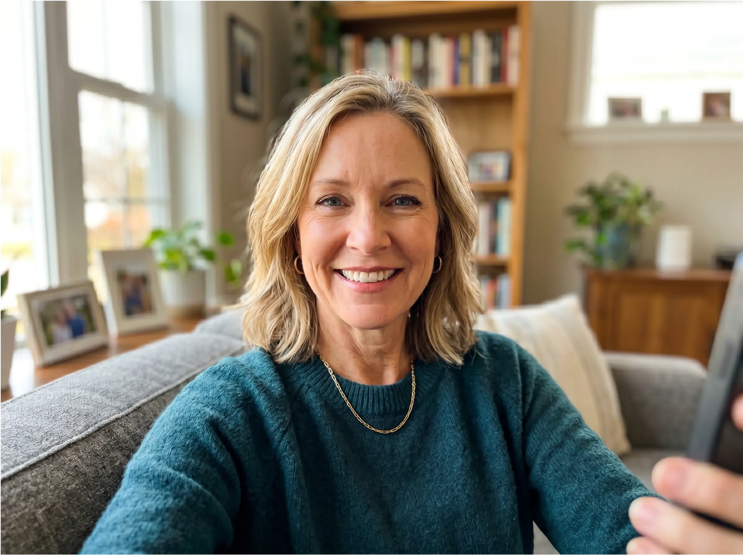 A smiling blonde woman in a teal sweater sits on a couch and looks at the camera.