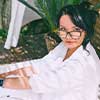 A person with dark hair and glasses, wearing a white shirt, sits and looks toward the camera.