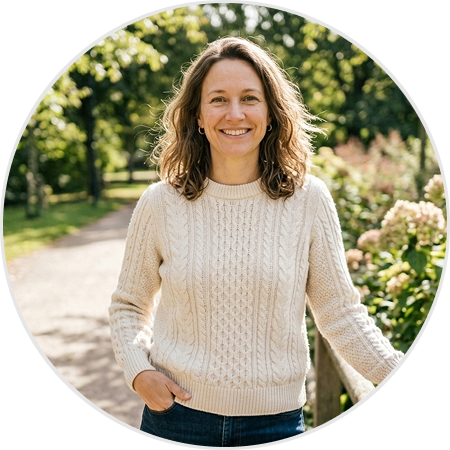 A smiling woman in a cream cable-knit sweater stands outdoors on a path in a park.