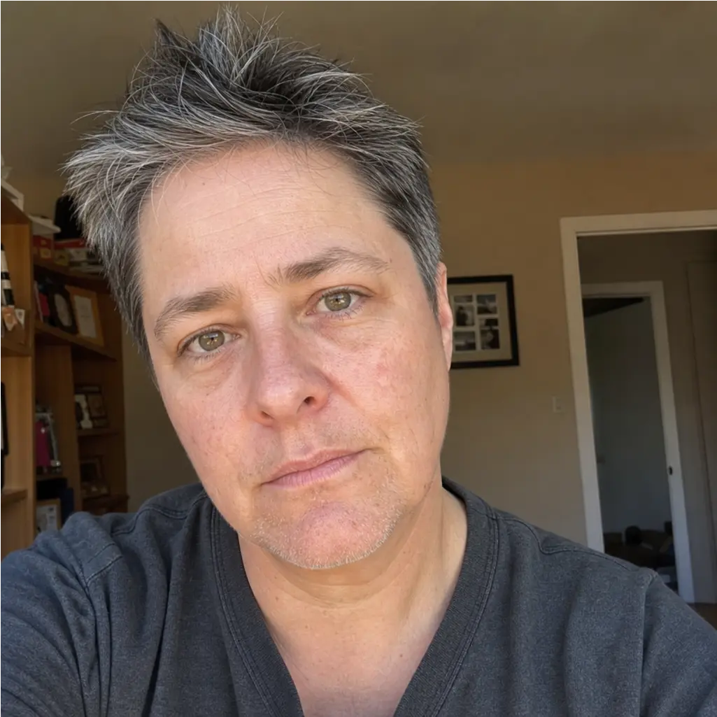 A close-up of a person with short, spiky salt-and-pepper hair and green eyes looking at the camera.