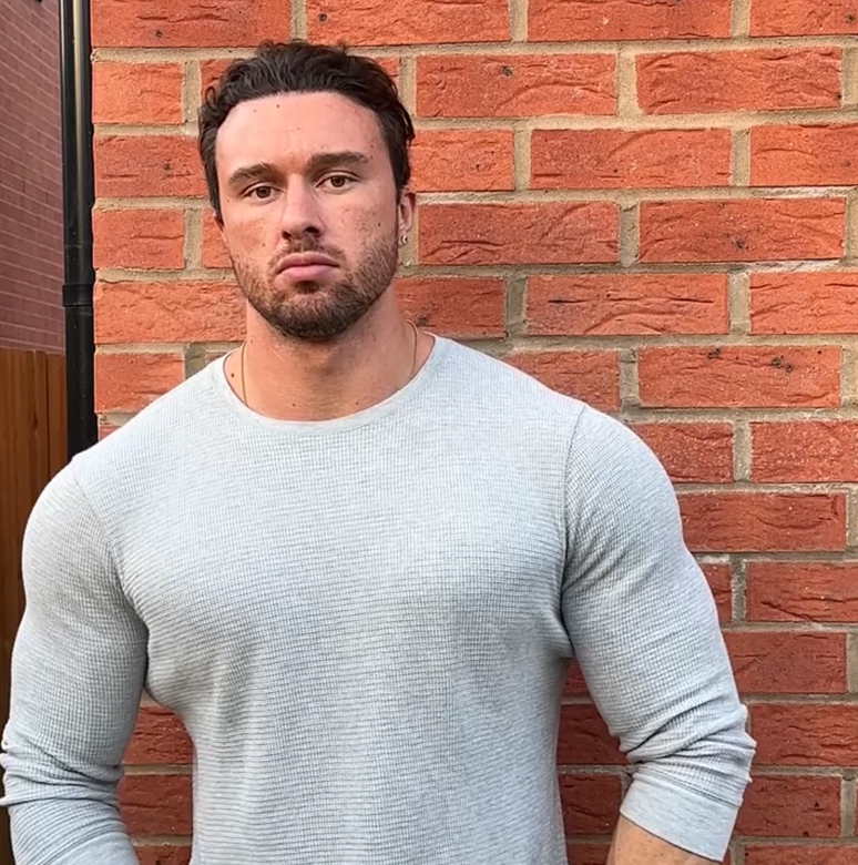 Man in a gray sweater standing against a red brick wall.
