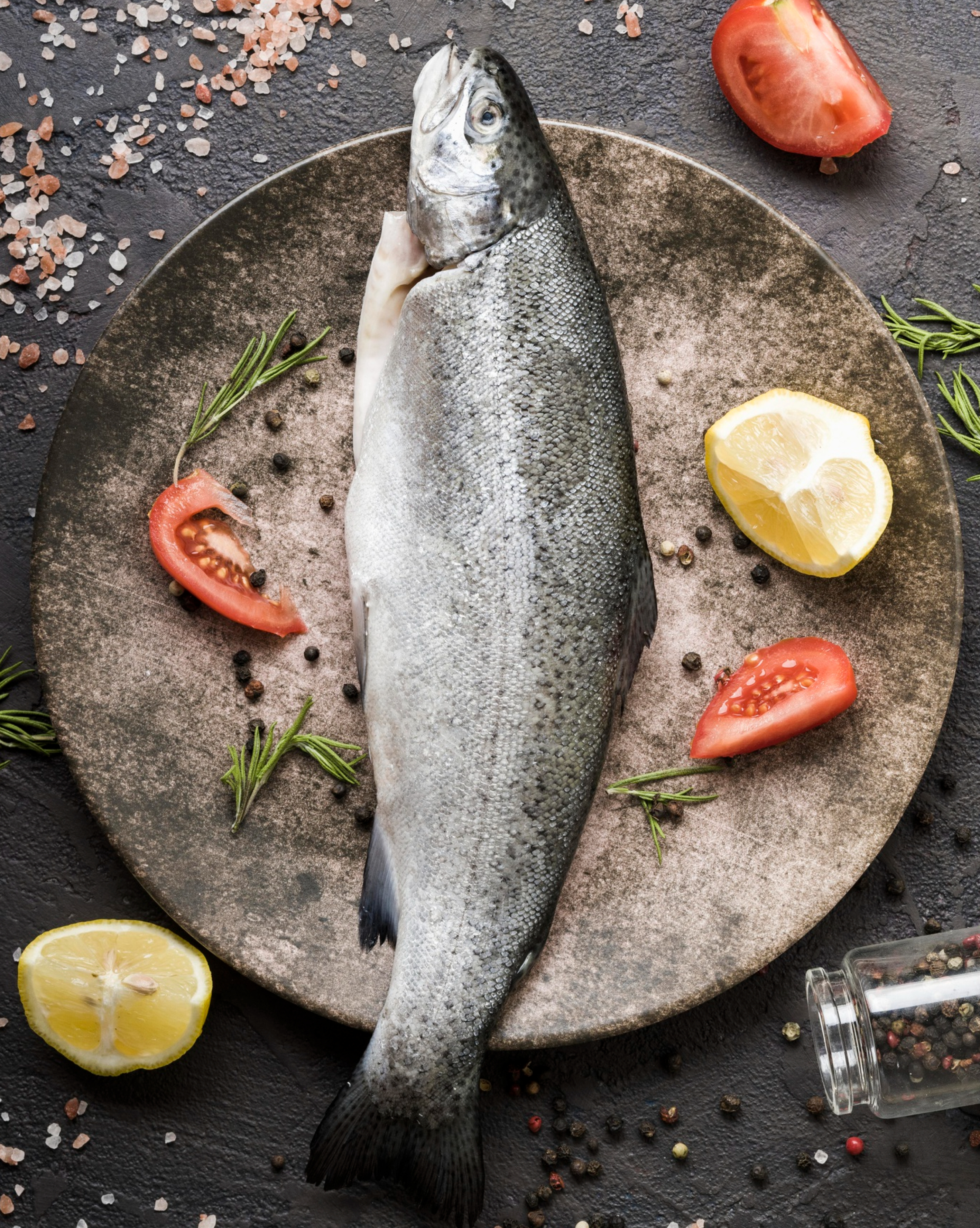 Whole fish on a plate with tomato, lemon, rosemary, peppercorns, and salt.
